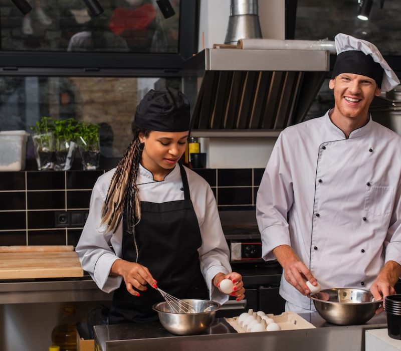 young african american sous chef in apron holding egg near bowl and whisk while cooking near happy